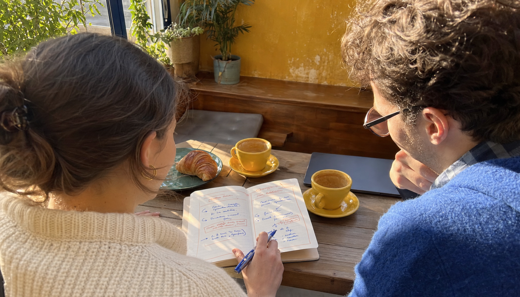 Deux personnes comparent deux options sur un carnet, à une table de café ensoleillée